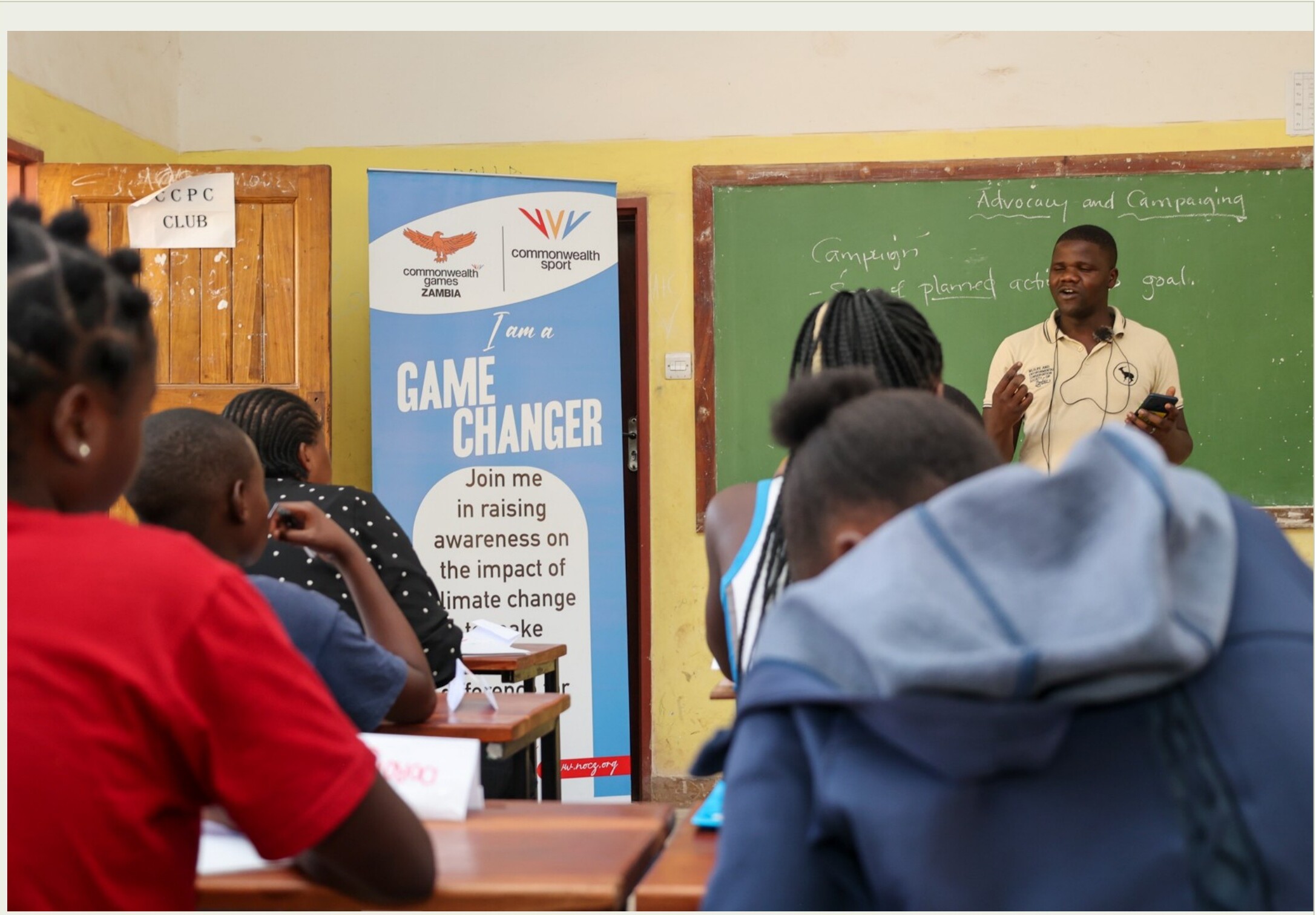 A facilitator delivering a Climate Game Changers advocacy and campaigning education session to students next to a Commonwealth Games Zambia banner